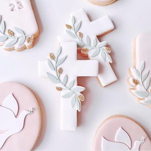 May include: White cross-shaped cookies decorated with light green leaves and gold accents. Other cookies include a pink circle with a white dove design and a rectangular cookie with the number 23.