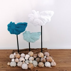 以下が含まれることがあります： ターコイズ、ブルー、ホワイトの3色のクロシェでできた貝殻が、小さな貝殻の山のある木製台座に飾られています。