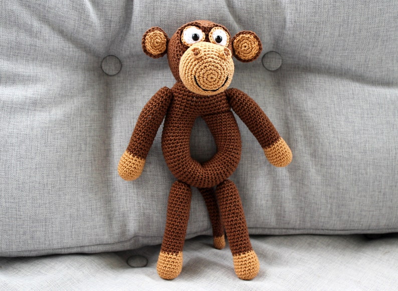 May include: A brown crocheted monkey toy with a ring for a body. The monkey has a smiling face with white eyes and a brown nose.