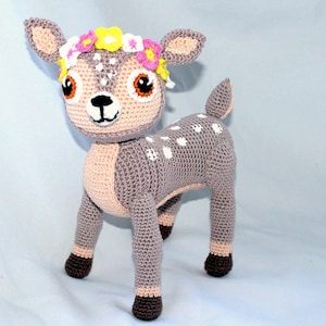 May include: A crocheted fawn wearing a flower crown. The fawn is grey with white spots and has brown hooves. The flower crown is pink, yellow and white.
