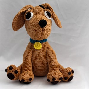 May include: A crocheted brown dog toy with black eyes, a black nose, and brown paws. The dog is wearing a blue collar with a yellow tag.