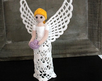 May include: A handmade crochet angel figurine with white wings and dress, holding a purple heart. The angel has yellow hair and a detailed, openwork design. The figurine is displayed on a dark surface.