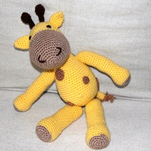 May include: A crocheted stuffed giraffe toy with brown spots and a brown nose. The giraffe is sitting on a white fabric surface.