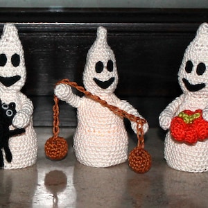 May include: Three crocheted ghost ornaments, each with a different accessory. One ghost holds a black spider, another holds a red apple, and the third holds a brown ball on a string.
