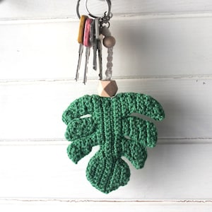 Puede incluir: Un llavero de hoja de monstera de crochet verde con una cuenta de madera y un juego de llaves.