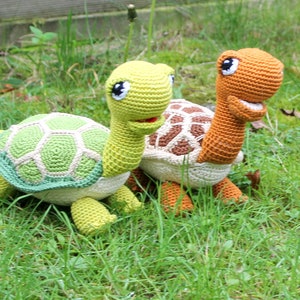 May include: Two crocheted turtle toys, one green and one orange, with smiling faces and detailed shells. The turtles are sitting on a bed of green grass.