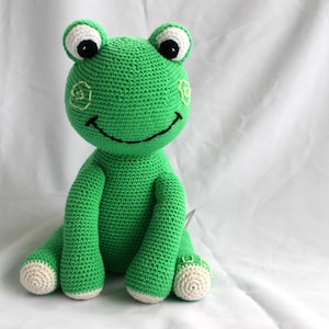May include: A green crocheted frog plush toy with black eyes, a white smile, and yellow embroidered details. The frog is sitting on a white surface.