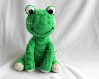 May include: A green crocheted frog plush toy with black eyes, a white smile, and yellow embroidered details. The frog is sitting on a white surface.
