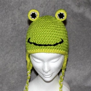 May include: Green crocheted frog hat with black eyes and a black smile. The hat has white and green yarn ties.