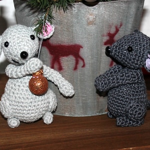 May include: Two crocheted mouse figurines. One is light gray and holding a small, glittery ornament. The other is dark gray. Both have black eyes and pink or purple inner ears. They are set against a rustic background.