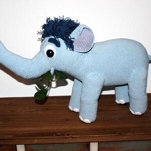 May include: A light blue crocheted elephant toy with a blue and white striped tail, a blue and white striped mane, and a large, open mouth. The elephant is holding a green plant in its trunk.