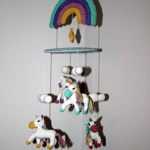 Può includere: Un mobile per neonati in crochet bianco con un arcobaleno, nuvole e quattro figure di unicorno. Gli unicorni hanno corpi bianchi, criniere arcobaleno e accenti verdi, blu e viola.