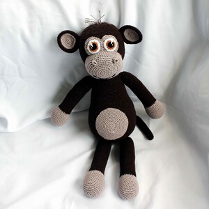 May include: A brown crocheted monkey toy with large brown eyes and a gray nose. The monkey is sitting on a white surface.