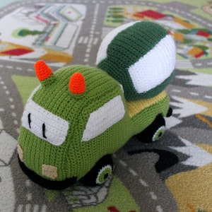 May include: A green crocheted toy cement mixer with a white and green crocheted cement drum. The toy is on a green and gray play mat.