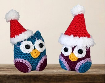 May include: Two small crocheted owl figurines, one blue and one purple, each wearing a red and white yarn Santa hat. The owls feature large white crocheted eye patches with black centres, yellow beaks, and colourful wing accents on their sides.