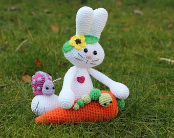 May include: A handmade crochet bunny with a sunflower crown and a red heart. A crochet snail and caterpillar sit on an orange carrot. The toys are white, pink, green, and orange, set against a green grass background. A whimsical, handcrafted Easter theme.