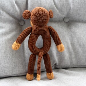 May include: A brown crocheted monkey toy with a ring in the middle of its body. The monkey has a long tail and brown yarn for its arms and legs.