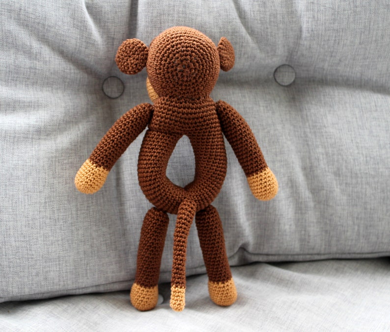 May include: A brown crocheted monkey toy with a ring in the middle of its body. The monkey has a long tail and brown yarn for its arms and legs.