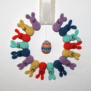 May include: A colorful wreath of crocheted bunnies with an Easter egg in the center. The bunnies are in various colors, including pink, purple, blue, green, yellow, and orange.