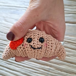 May include: A small, light brown crocheted croissant toy featuring two black bead eyes, a stitched dark blue smile, pink embroidered cheeks, and a bright orange accent on one side. The item is crafted with visible yarn stitches.