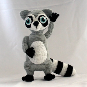 May include: A gray and white crocheted raccoon plush toy with black eyes and a black and white striped tail. The raccoon is standing on a white surface and has its right arm raised.