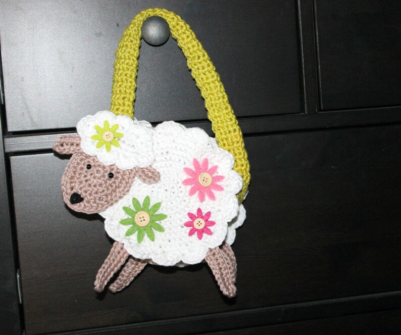 May include: A white crocheted lamb shaped bag with a green strap and pink and yellow flower accents.