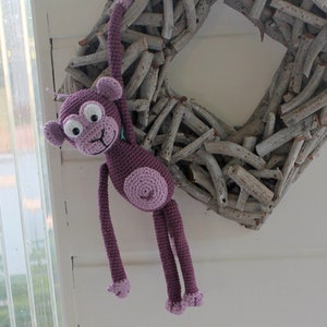 Puede incluir: Un juguete de mono de crochet morado colgando de una corona de ramas. El mono tiene una cola larga y está colgando boca abajo.