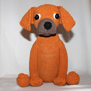 May include: A crocheted orange dog toy with black eyes and a brown nose. The dog is sitting with its legs crossed.