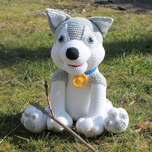 May include: A crocheted husky dog toy with gray and white fur, blue eyes, and a blue and yellow collar. The toy is sitting on a bed of green grass.