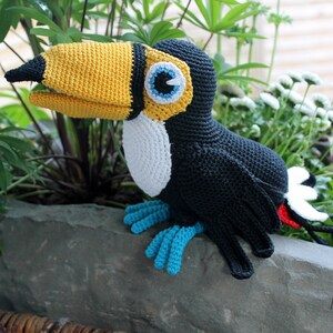 May include: A crocheted toucan plush toy with a yellow beak, black body, white accents, and blue feet. The toucan is sitting on a gray stone surface.