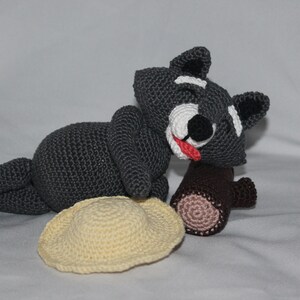 May include: A gray crocheted raccoon plush toy with a red tongue sticking out, lying on its side with a brown log and a yellow crocheted hat.