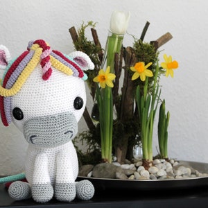 Può includere: Un unicorno bianco all'uncinetto con una criniera e una coda arcobaleno. L'unicorno è seduto su una superficie nera davanti a un piccolo arrangiamento di narcisi gialli e un tulipano bianco.