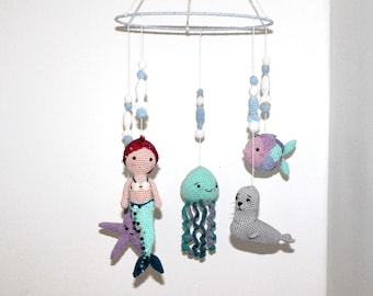 May include: A handmade crochet baby mobile featuring sea creatures. The mobile includes a mermaid, jellyfish, fish, and seal, all in soft pastel colours. The mobile is suspended from a white and blue ring with decorative beads.