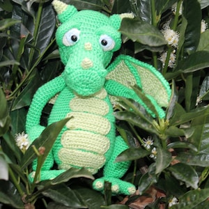 May include: A green crocheted dragon toy with large eyes and a light green belly. The dragon is sitting in a bush with green leaves and white flowers.