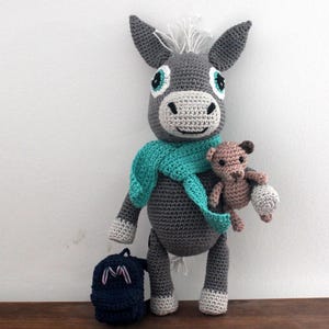 May include: A gray crocheted donkey toy with teal blue scarf and a brown crocheted teddy bear in its arms. The donkey has white fur on its head and large blue eyes. The donkey is holding a small blue backpack with the letter "M" on it.