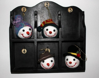 May include: A black wooden shelf with six compartments, each with a number from 1 to 6. Each compartment holds a crocheted snowman ornament with a different hat and facial expression.