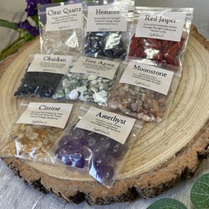 Könnte beinhalten: Sortiment kleiner, polierter Edelsteine in durchsichtigen Plastikbeuteln, darunter Bergkristall, Hämatit, Roter Jaspis, Obsidian, Baumachat, Mondstein, Citrin und Amethyst. Jeder Beutel hat ein Etikett mit dem Namen und den Eigenschaften des Edelsteins.