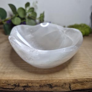 May include: A heart-shaped, translucent white bowl. The bowl is made of a crystalline material and sits on a natural wood surface. The bowl's shape is organic, with smooth curves and a polished finish. The background includes greenery.