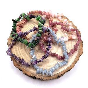 May include: A selection of gemstone chip bracelets in shades of green, pink, purple, and blue. The bracelets are presented on a natural wooden slice, highlighting the diverse colours and textures of the stones. The bracelets are composed of small, irregularly shaped chips.