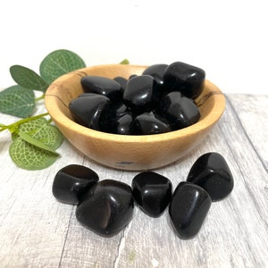May include: A wooden bowl filled with black obsidian tumbled stones. The bowl is sitting on a light grey wooden surface. There are also several black obsidian tumbled stones outside of the bowl.