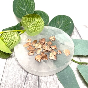 May include: A collection of small, irregular pieces of copper metal, with a rough, textured surface, arranged on a white, circular surface. The copper pieces are a warm, reddish-brown colour.