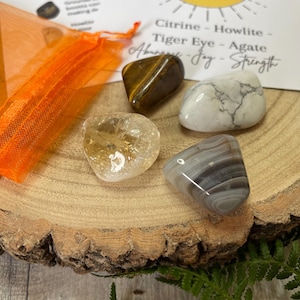 May include: Four polished gemstones: citrine, tiger eye, agate, and howlite, displayed on a wooden slice. An orange mesh bag and a card with text are in the background. The card lists the stones and their associated qualities.