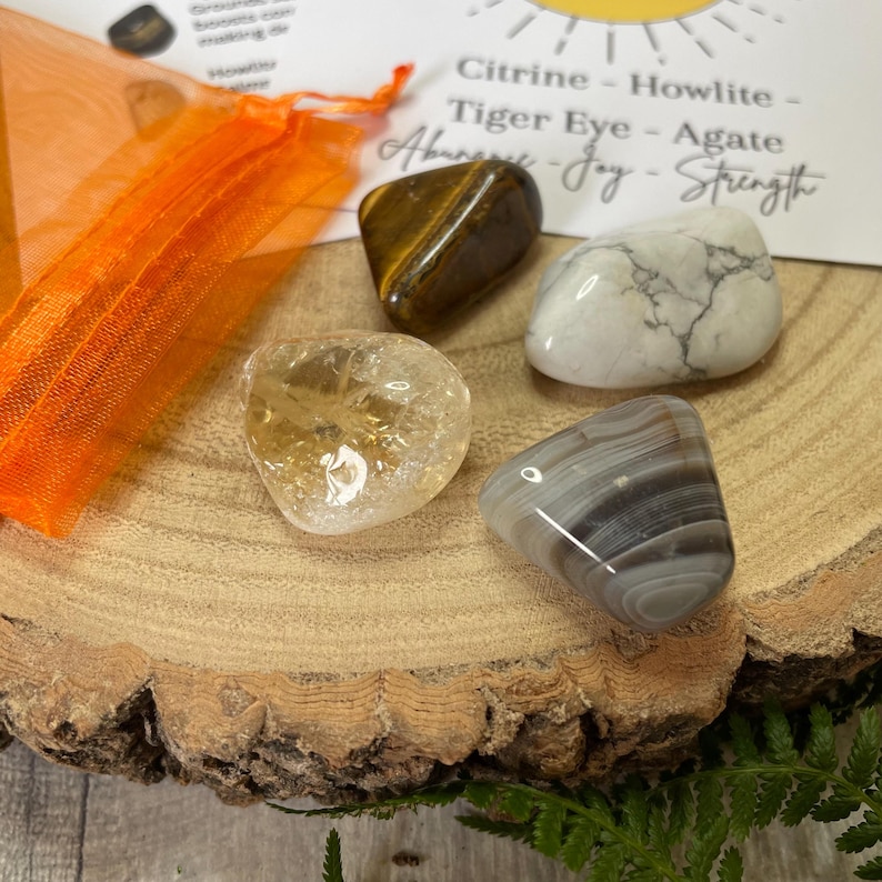 May include: Four polished gemstones: citrine, tiger eye, agate, and howlite, displayed on a wooden slice. An orange mesh bag and a card with text are in the background. The card lists the stones and their associated qualities.