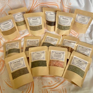 May include: A collection of spice packets, each labelled with the spice name in a decorative frame. Spices include cinnamon, fennel, aniseed, cloves, rosemary, garlic, dill seed, black salt, thyme, mint, pink salt, bay leaf, and sage. The packets are beige with a clear window.