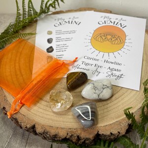 May include: A collection of gemstones, including citrine, tiger eye, and agate, displayed on a wooden surface. An orange organza bag and a Gemini zodiac information card are also present. The card lists the gemstones and their associated qualities.
