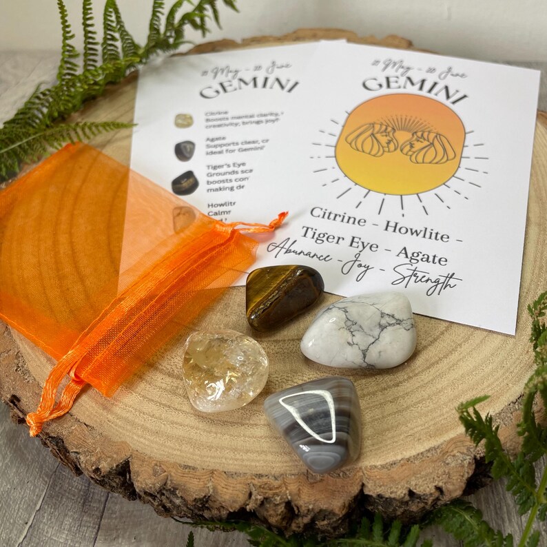 May include: A collection of gemstones, including citrine, tiger eye, and agate, displayed on a wooden surface. An orange organza bag and a Gemini zodiac information card are also present. The card lists the gemstones and their associated qualities.