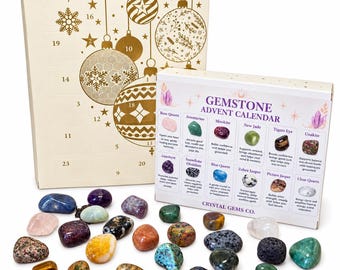 Op de afbeelding: Een edelsteen adventskalender set met een grote crèmekleurige doos met gouden kerstornamenten en een kleinere witte doos met het label Crystal Gems Co. die verschillende stenen zoals rozenkwarts, amethist en tijgeroog toont met losse gepolijste kristallen.