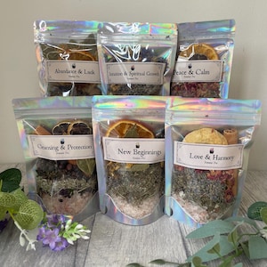 May include: Six iridescent pouches filled with dried herbs, flowers, and citrus slices. Each pouch has a label with text such as "Abundance & Luck" and "New Beginnings". The pouches are arranged on a light-colored surface, with greenery in the foreground.