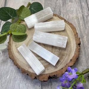 Peut inclure: Cinq bâtonnets de sélénite blanche sur une surface en bois. Les bâtonnets sont lisses et ont une texture naturelle striée.