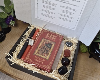 May include: A Requiem Arcana Tarot Pack in a black box, including a deck of 78 tarot cards, two candles, crystals, and a card with text. The tarot card box is red with gold accents.